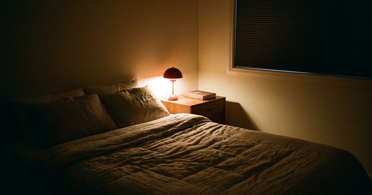 Dark bedroom lit only by a small warm red lamp on a nightstand beside a made bed