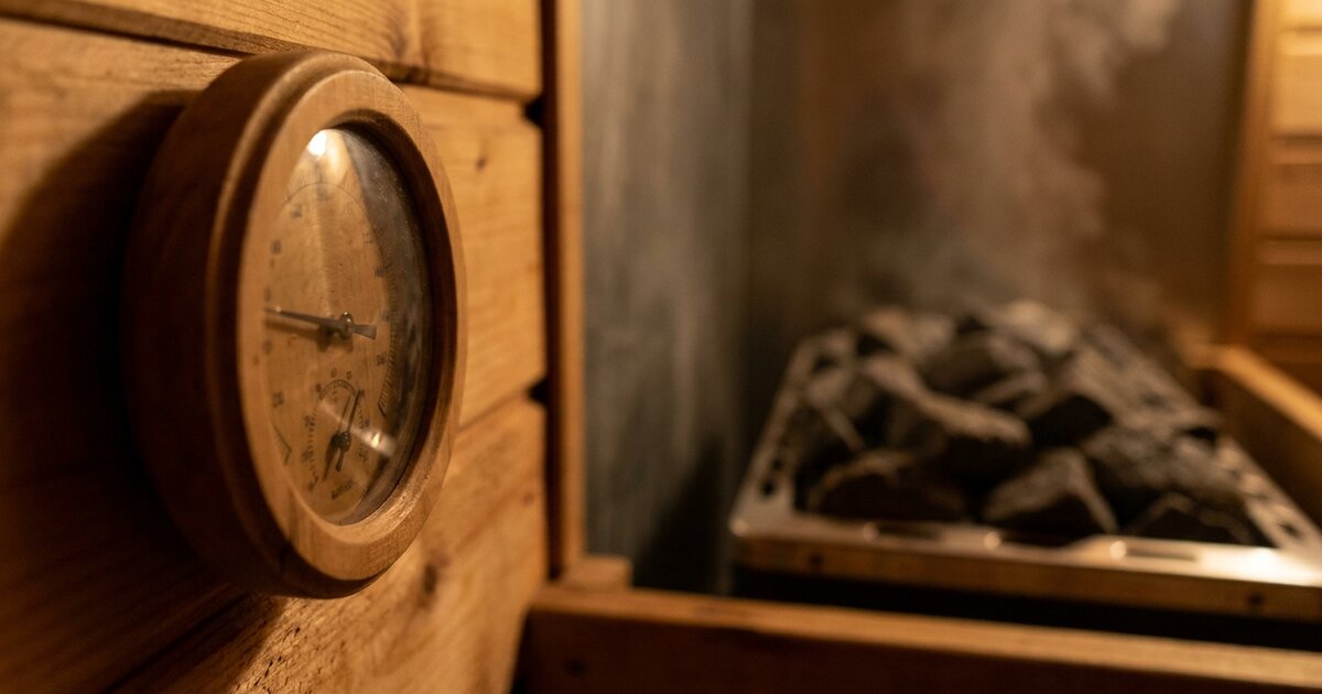 Round analog thermometer on a cedar wall inside a traditional sauna with hot stones behind it